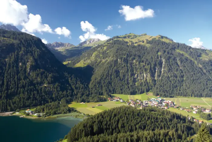 Landschaft mit See und einem kleinen Dorf und Bergen im Hintergrund.